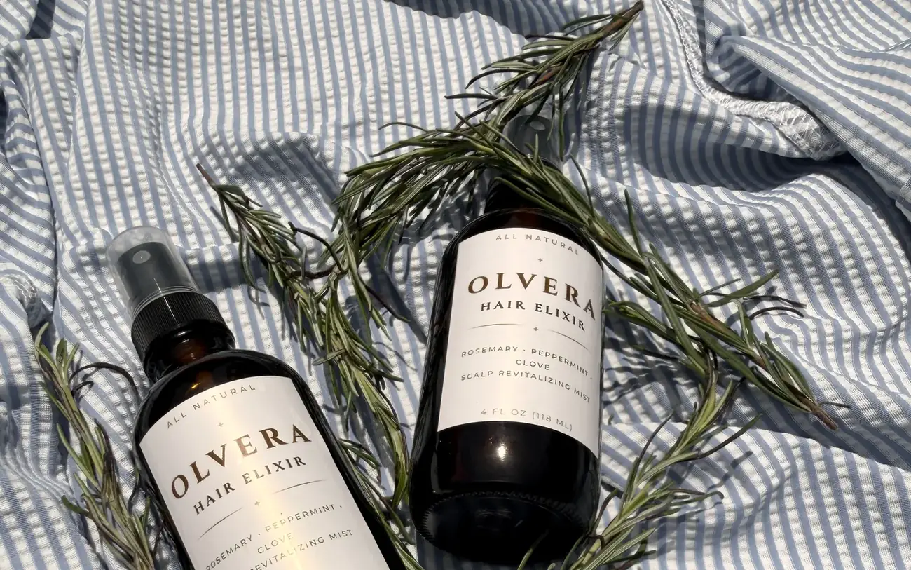 Two bottles of OLVERA Hair Elixir surrounded by fresh rosemary sprigs